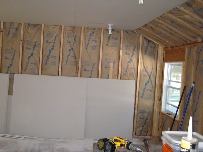 A studded wall with the paper side of fiberglass batts showing with drywall leaning against the wall. There are tools all around the room.