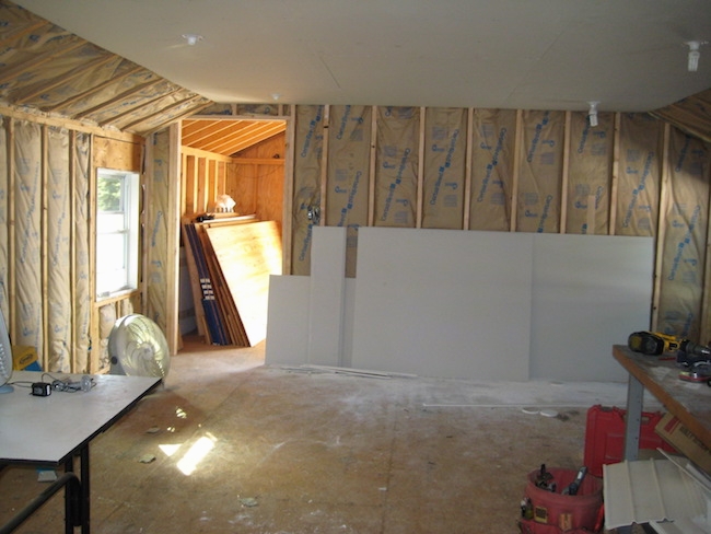 A studded room with fiberglass batts lining the walls and drywall leaning against the wall. There is a fan in the room and a white table.
