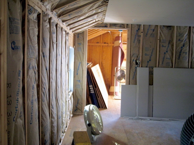 A room lined with fiberglass batts with drywall leaning against the wall that has not been put up yet. There is a fan sitting on the plywood floor.