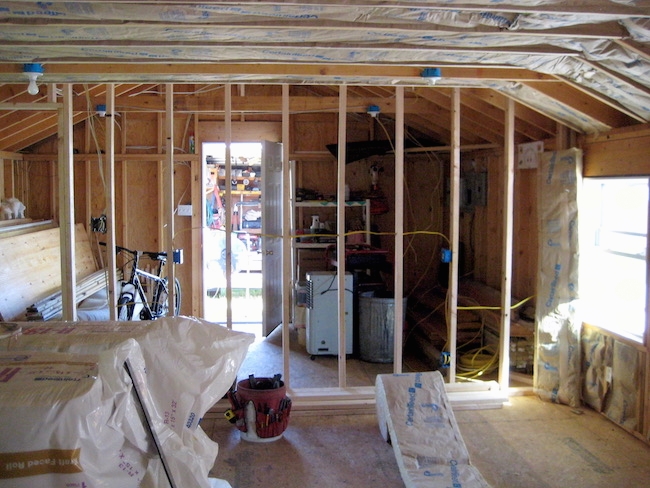 A room with the studs showing and fiberglass batts lining the walls and rolls of the spun glass on the floor awaiting instulation. There is a kids bicycle in the back of the room.