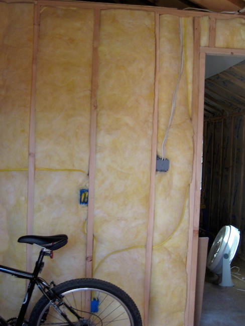 A room with yellow fiberglass in an open wall with electrical outlets and wires with a black bicycle parked in front of it. There is a white fan on the floor in the other room.