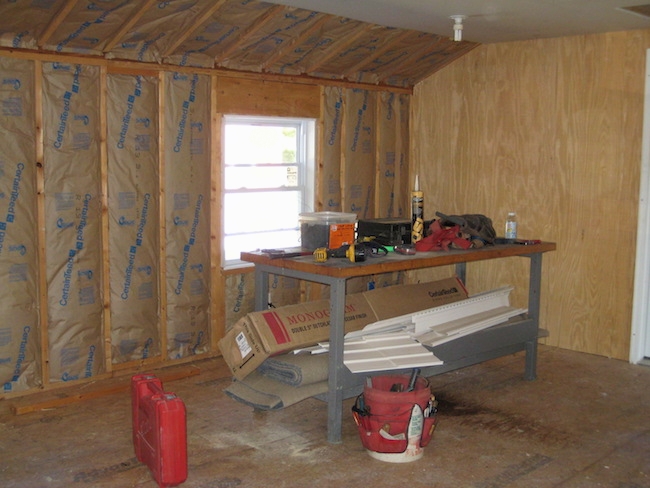 A room being built with fiberglass batts lining the studded wall up to the ceiling where the drywall was put up. There is a work bench and tools in front of the wall.