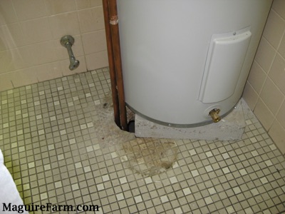 A hot water heater on top of a tiled floor with a hole in the floor where the pipe leaves the heater.