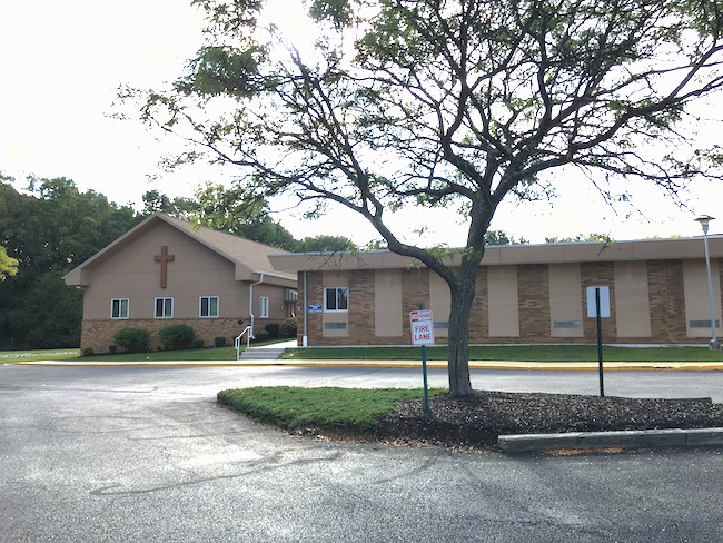 A brown one story building with a cross on the front. View from the parking lot showing a turn around area and a fire lane.