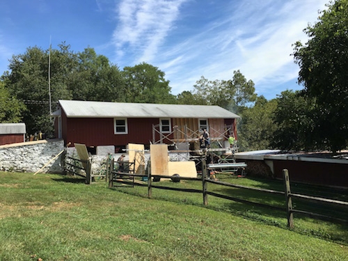 A distant view of a red barn with scaffolding and workers removing fiberlass insluation. There is a white stone wall and a wooden split rail fence.