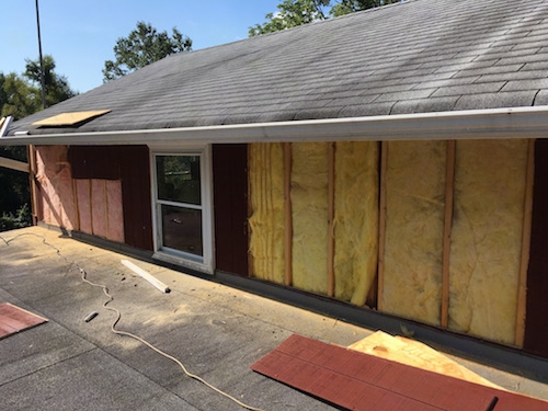 A wall of a red barn that has a roof coming off of the lower area with pink and yellow batts of fiberglass exposed in the walls after the T1-11 was removed.