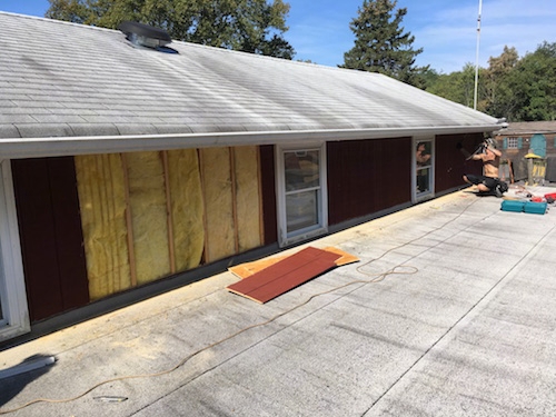 The wall of a barn with a construction worker removing the T1-11 to get the fiberglass batts out. There are yellow batts showing where it was already removed.