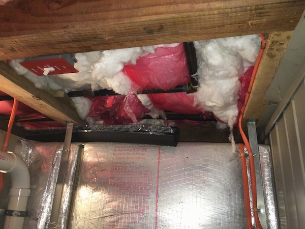 The ceiling on the lower level of a shore house with fiberlglass insulation hanging down exposed to the air where the pink plastic is ripped. There are wires and pipes around it.