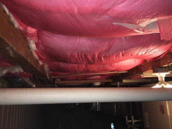 The ceiling at a beach house on the lower level showing pink plastic holding up fiberglass insulation which is attached to the ceiling. The plastic is ripping and the insulation is exposed