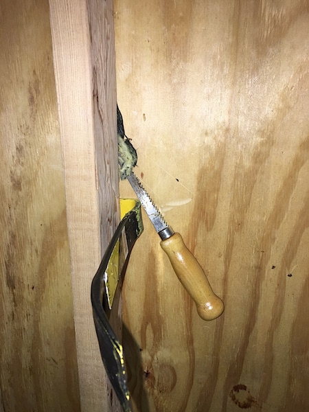 A crow bar and a saw prying out yellow fiberglass insulation that is stuck to black paper from behind a beam in a studded wall.