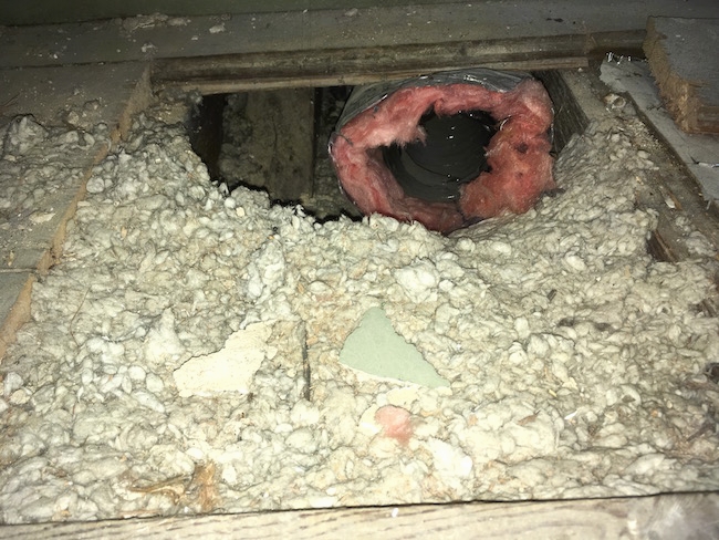 A square area on the floor where boards were removed with gray blown-in insulation exposed with a black flex duct with pink fiberglass around it poking out.