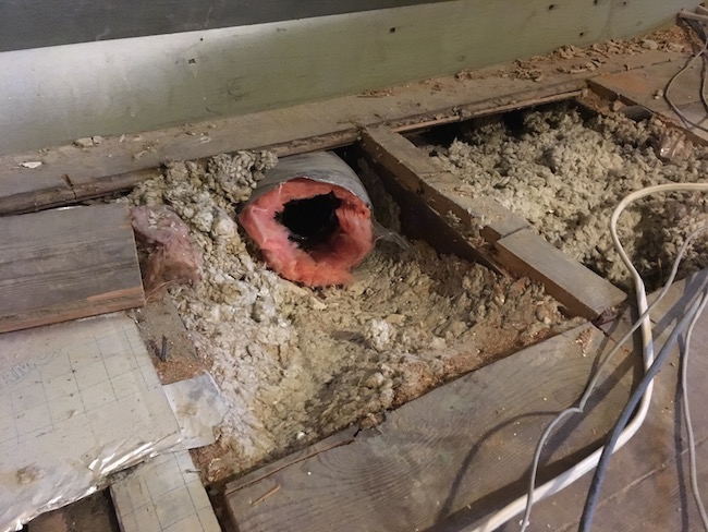 Boards removed from an attic floor showing the loose blown in fiberglass with a flex duct poking out and wires off to the right side.