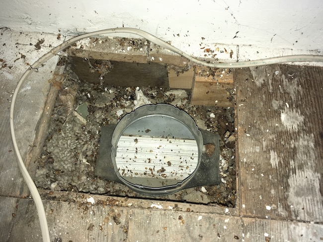 Looking down into an attic floor that had a floor board removed exposing a very dirty area of blown in fiberglass and a hard silver duct.