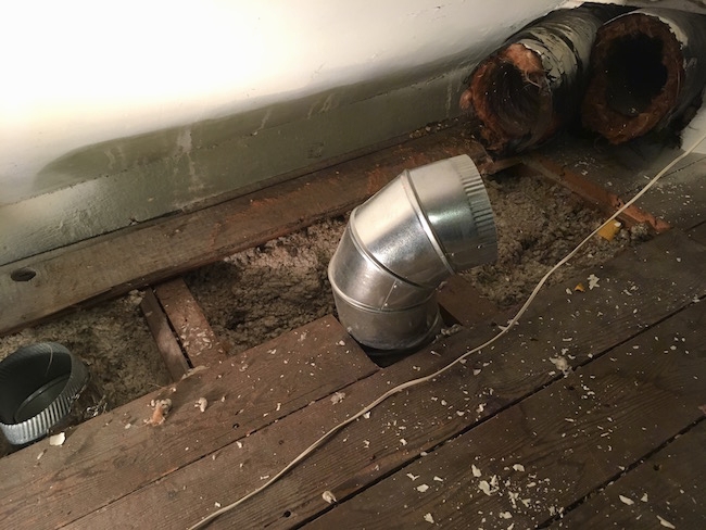 Flex ducts poking out from the wall in an attic with some boards removed with two silver ducts also sticking out of the floor.