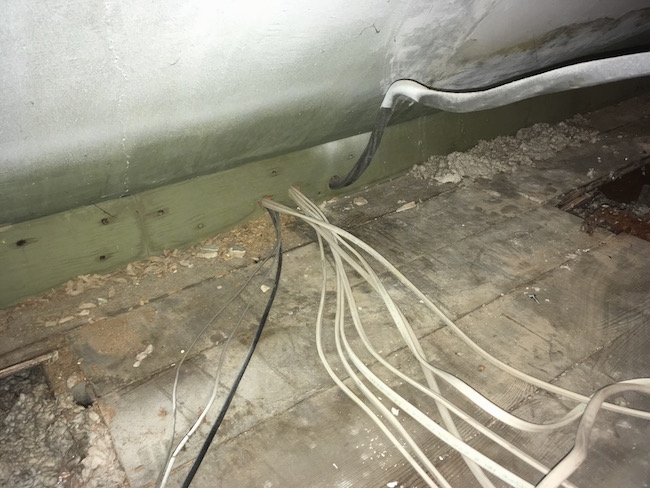 An attic floor with white wires running into the wall and gray blown in fiberglass sprinkled about with two floor boards removed.