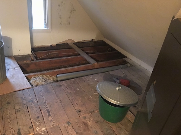 An attic floor with the boards removed towards the back showing the studs and some gray fiberglass insulation and a green trash bucket with a silver trash can lid laying on top of it and a long skinny window with the sun shining in.