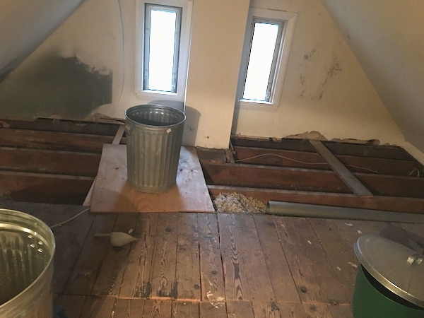 An attic floor with the boards removed towards the back of the room with two long skinny windows with the sun shining in, two medal trash cans and a green bucket with a silver trash can lid on top of it. You can see the blown in gray fiberglass insulation between one of the grooves.