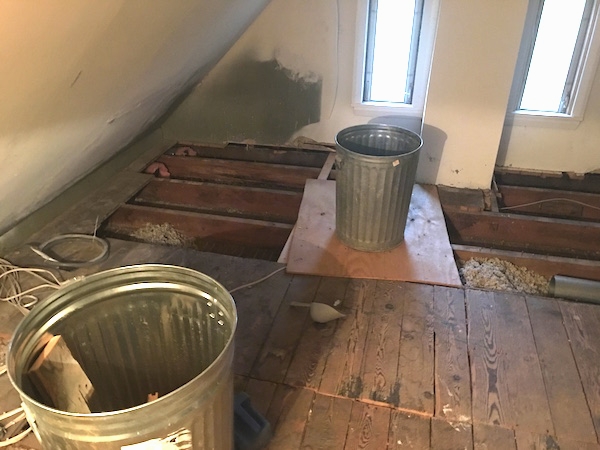 An attic floor with the boards removed towards the back of the room with two long skinny windows with the sun shining in and two medal trash cans and white wires. You can see the blown in gray fiberglass insulation between one of the grooves.