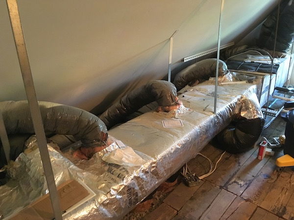 An AHVC system up in an attic with a long insulated metal duct running out of it and four black flex ducts connected to the long run. There is exposed fiberglass at the base of the flex ducts.