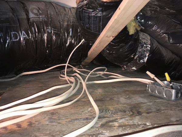 Large black flex ducts on a wooden floor with white wires going down into the floor, exposed yellow insulation and a 2 x 4 piece of wood holding up one of the ducts.