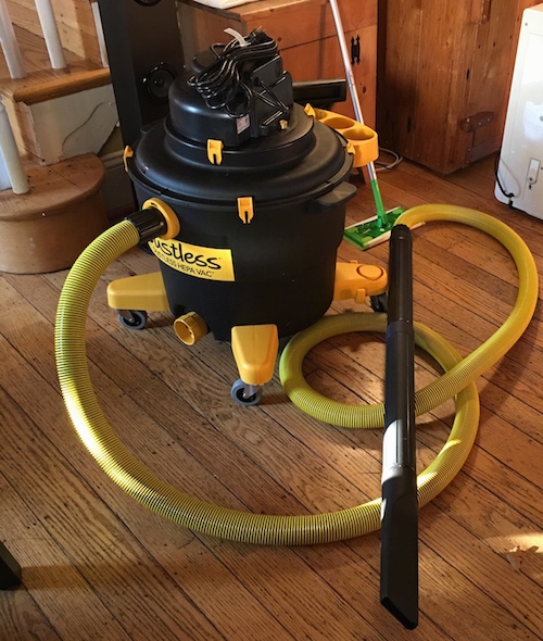 A black and yellow shop vac sitting on a hardwood floor in front of a set of stairs. There are two wooden cabents and a swiffer mop behind it.