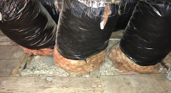 A hole in a wooden floor with large black flex ducts going down into the floor. There is orange fiberglass at the base of the ducts hanging out from the black plastic cover and loose gray fiberglass around the bottom of them on the floor.