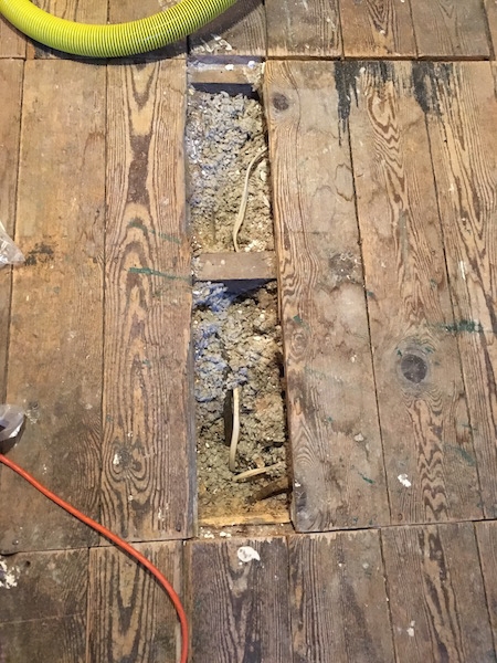 A wooden attic floor with one board removed exposing the blown in fiberglass insulation. You can see a wire running inside of the fiberglass. There is a yellow shop vac tube and an orange extension cord on each side of the hole in the floor.