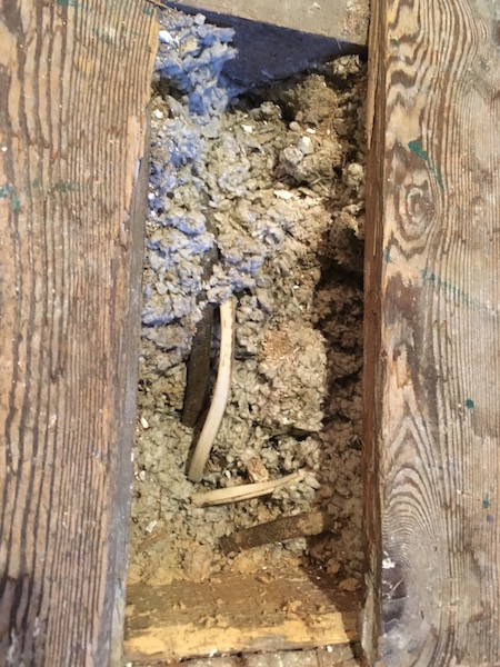 A wooden attic floor with one board removed exposing the blown in fiberglass insulation with a white wire running through it.