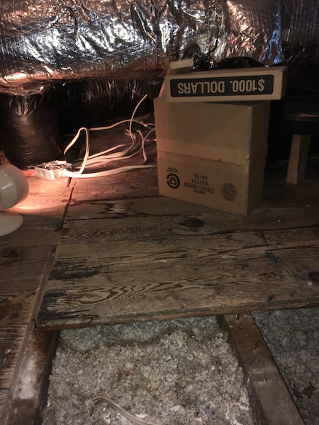 An attic with flex ducts above and near the wall, a wooden floor with some boards removed showing blown in fiberglass insulation and some cardboard boxes.