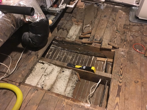 A view looking down at a wooden floor with floor boards removed showing thick white blown in fiberglass in the floor cavity. There is a cental air unit to the left