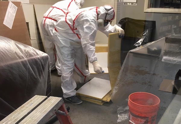 Two men wearing hazmat suits, respirators and gloves inside of an offfice that has plastic covering up all of the office furniture. The first man is picking up one of the many fiberglass ceiling panels.