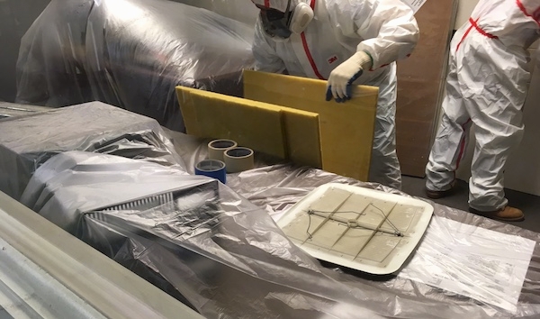 A man wearing a hazmat suit and a respirator with his gloved hand on a yellow fiberglass ceiling panel with the desk inside the office covered in plastic. A second suited worker is behind him.