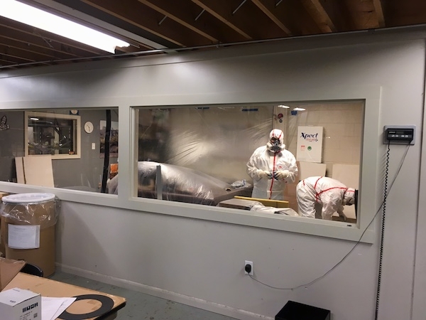 Two men wearing hazmat suits, respirators and gloves inside of an offfice that has plastic covering up all of the office furniture. There are fiberglass ceiling panels around them.