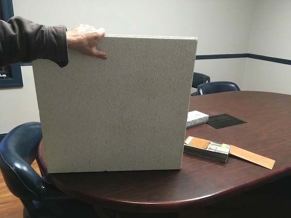 A man's hand holding a square white ceiling tile up on a brown desk inside of an office.