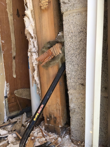 A crow bar in the end of a cinder block wall inside of a building pulling out a clump of moldy fiberglass sticking out from between the cinder blocks and the wood frame.