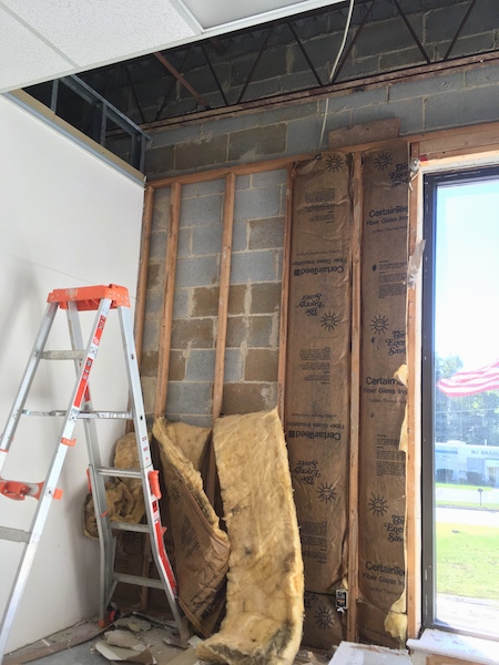 A cinder block wall with yellow fiberglass batts hanging out from between the wood frame work. There is a ladder in front of it and an American flag blowing in the wind outside the front glass door.