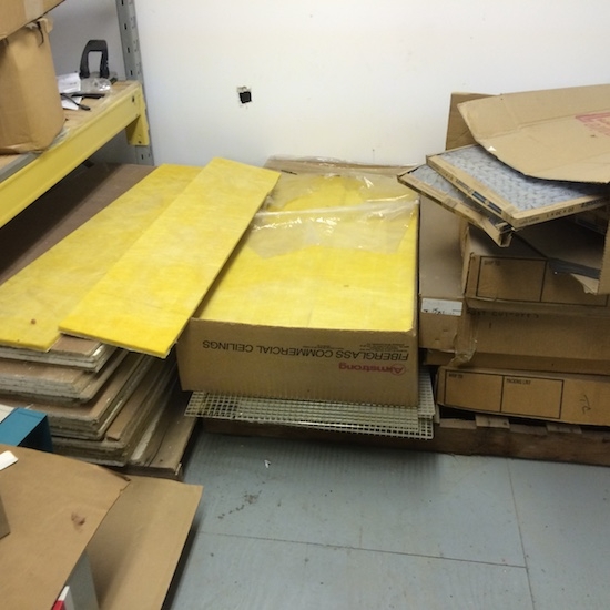 Piles of yellow fiberglass commercial ceiling panels in brown cardboard boxes sitting on top of a gray floor behind a white wall that has a hole in the drywall.