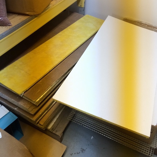 Stacks of yellow fiberglass commercial ceiling panels in brown cardboard boxes sitting on top of a gray floor in front of a white wall.