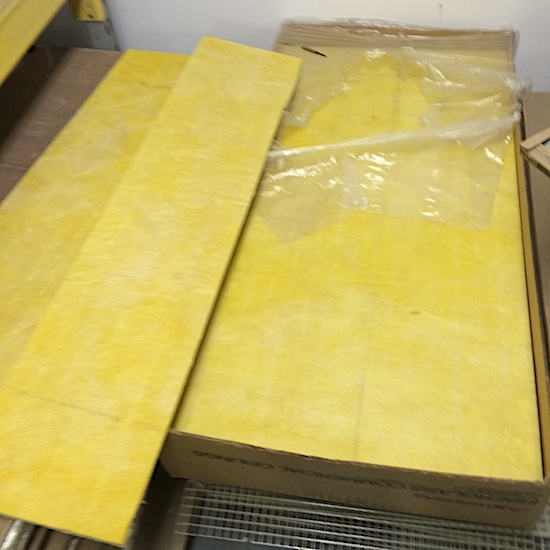 Two piles of yellow fiberglass commercial ceiling panels in brown cardboard boxes sitting on top of a gray floor in front of a white wall.