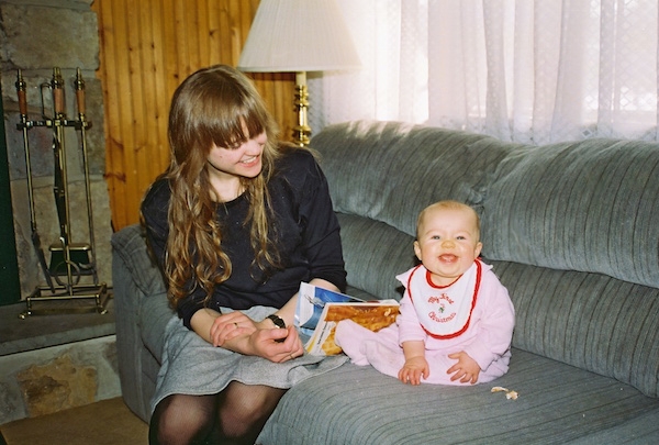 A baby sitting on a couch smiling at the camera as her mother sits next to her and smiles down at her