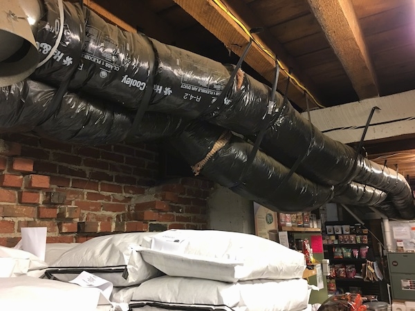 Two big black round flex ducts running across the length of a feed store with white horse feed bags stacked under them. The ducts have exposed fiberglass showing in the seams.