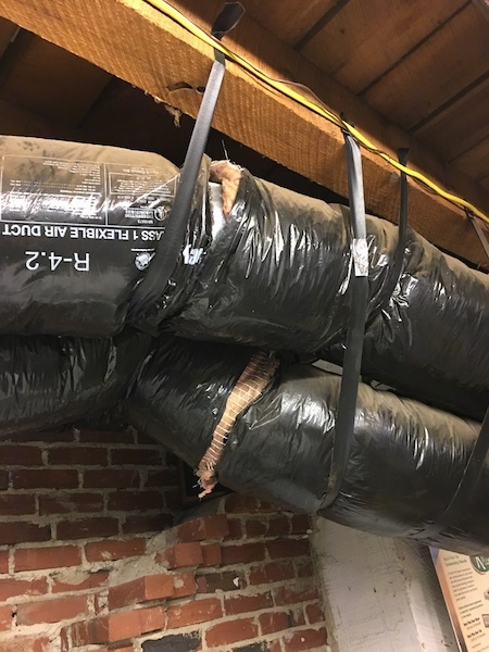 Close up - Two big black round flex ducts running across the length of a feed store. The ducts have exposed fiberglass showing in the seams. There is a brick wall behind them.