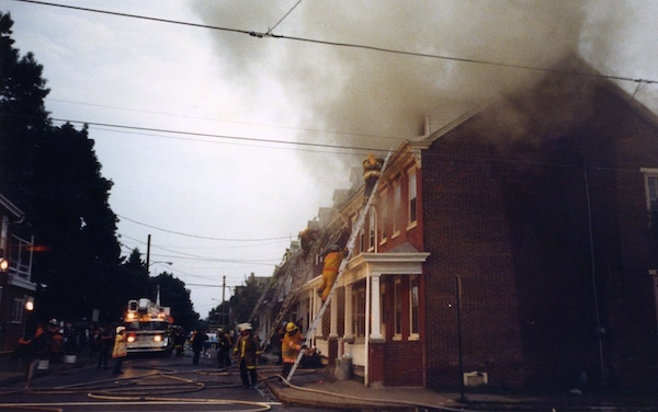 A street in a town of row homes with smoke coming from the end house. There are fire fighters with a ladder against the house.