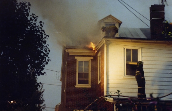 The second floor of a house with flames coming from the roof and a couple of firemen on the roof. There is smoke coming from the house.