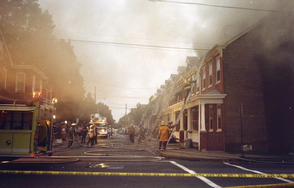 The view down a street with smoke coming from one of the homes, fire trucks and firemen in street and ladders leaning on the houses in various places.