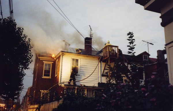 The second floor of a brick and white house with fire fighters on the roof. There is smoke coming from the house.