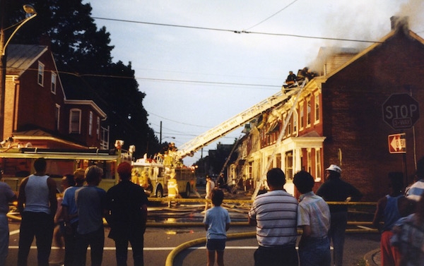 A street in a town of row homes with smoke coming from the end house. There are fire fighters with a ladder against the house spraying water on it and a crowd of people watching from the street.