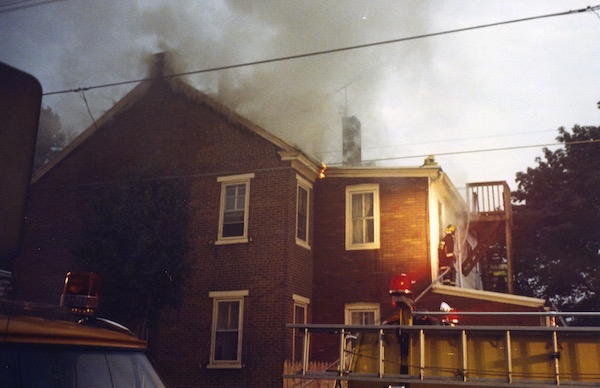 A brick house with smoke coming from it and fire fighters walking on the roof.