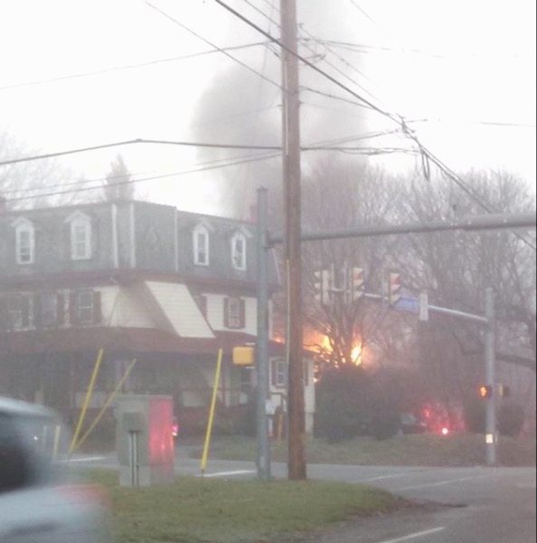 A three story tan and red home with a house burning behind it.