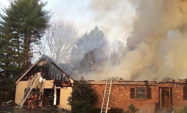 A burning house with a ladder leaning on the side of it, the roof gone and smoke coming from it.